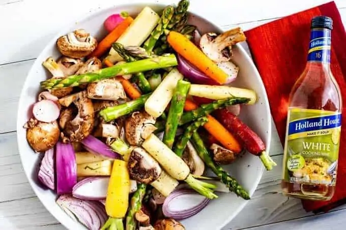 White bowl with asparagus, carrots, mushrooms, onions, and radishes mixed together and a bottle of white cooking wine sitting next to it.