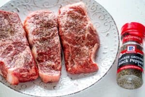 Photo of steaks seasoning with salt and pepper on a white plate.