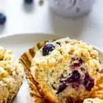 Close up photo of a blueberry quinoa muffin with two other muffins beside it sitting on a white plate with a white mug behind it.