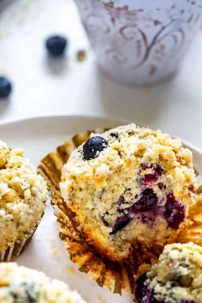 Close up photo of a blueberry quinoa muffin with two other muffins beside it sitting on a white plate with a white mug behind it. How To Make Blueberry Quinoa Muffins (recipe)