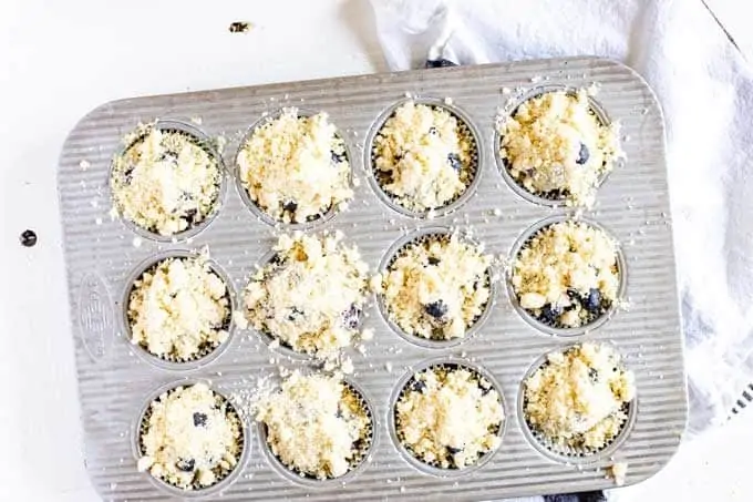 Photo of a muffin tin with blueberry quinoa muffin batter that has been topped with a crumbly topping.