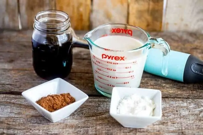 Photo of a mason jar of cooled coffee, a measuring cup with milk, and white prep bowls of cocoa powder and Swerve sweetener with an immersion blender behind it.