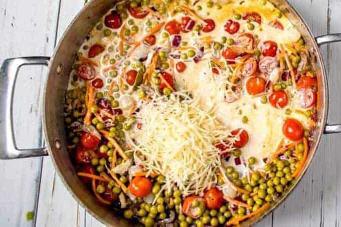 Photo of half and half and cheese being added to a large skillet with cooked baby peas, carrots and tomato.