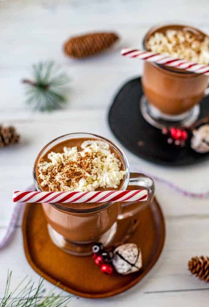Photo of two glasses slow cooker hot chocolate on wodden plates with holiday decor surrounding them.