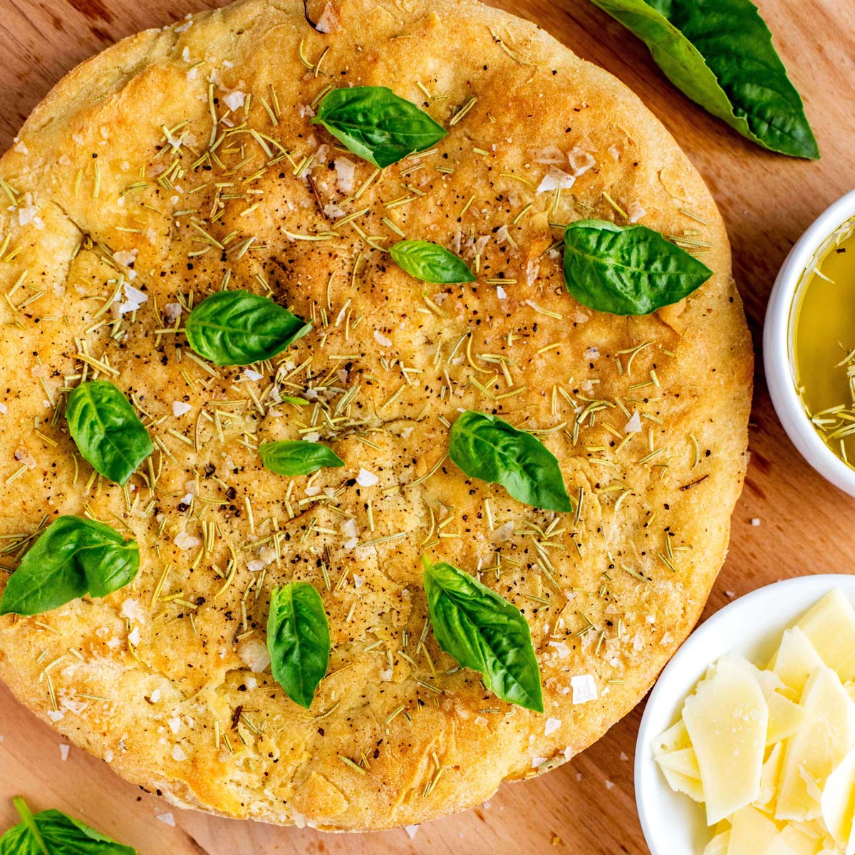 Square Photo of Gluten Free Rosemary Focaccia Bread on a cutting board garnished with basil and surrounded by olive oil and parmesan cheese.