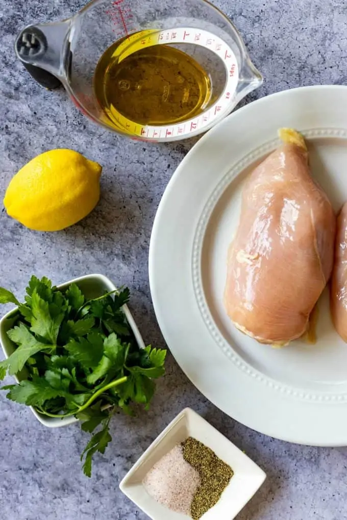Photo of chicken breast on a white plate, oil, lemon, parsley, and seasonings.