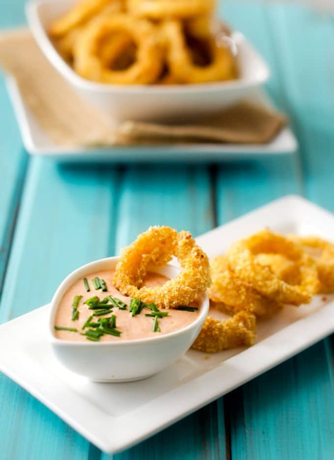 Photo of a healthy onion ring being dipped into a dipping sauce with a bowl of onion rings behind it.