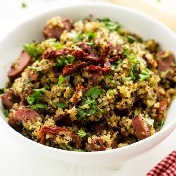 Square close up photo of a white bowl with quinoa and sausage garnished with parsley and sun dried tomatoes.