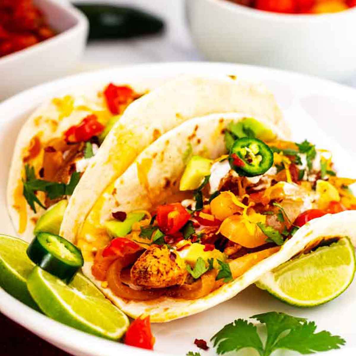 Square photo of two slow cooker chicken fajitas on a white plate garnished with cilantro and jalapeno.