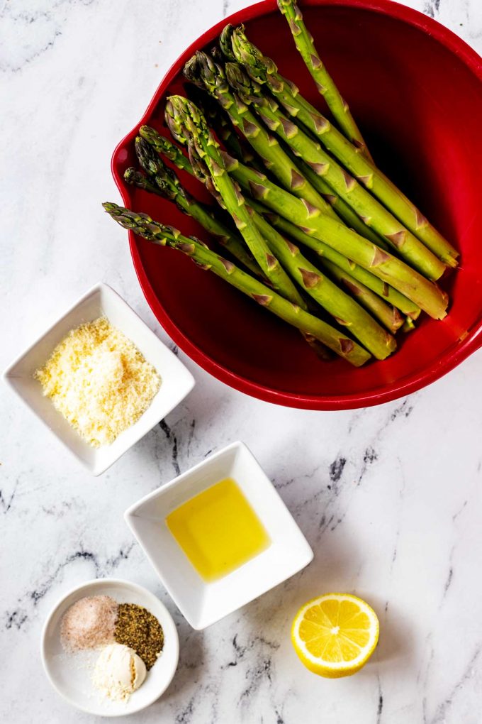 Photo of a bowl of asparagus, a half lemon and small prep bowls with parmesan, avocado oil, and seasonings.