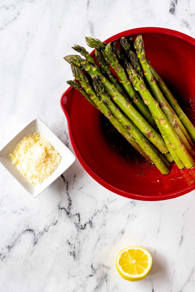 Bowl of seasoned asparagus sitting next to a small dish of parmesan and a half of lemon.
