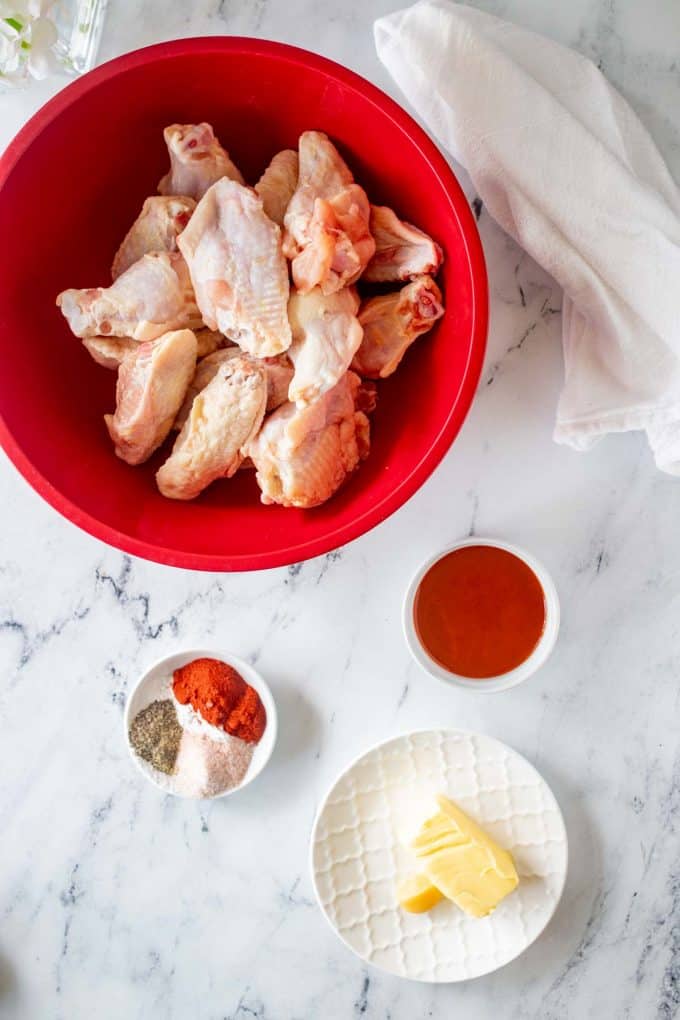 A bowl of chicken wings with small prep containers of buffalo sauce, seasonings, butter, and garlic next to it.