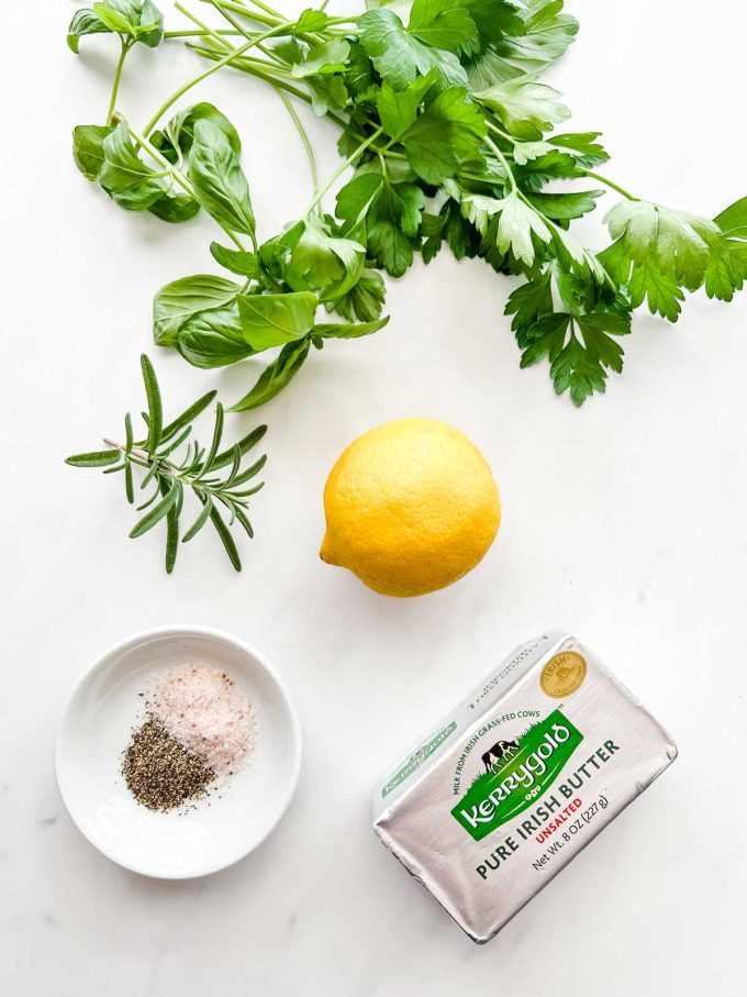 Photo of herbs, butter, seasonings, and lemon on a white background.