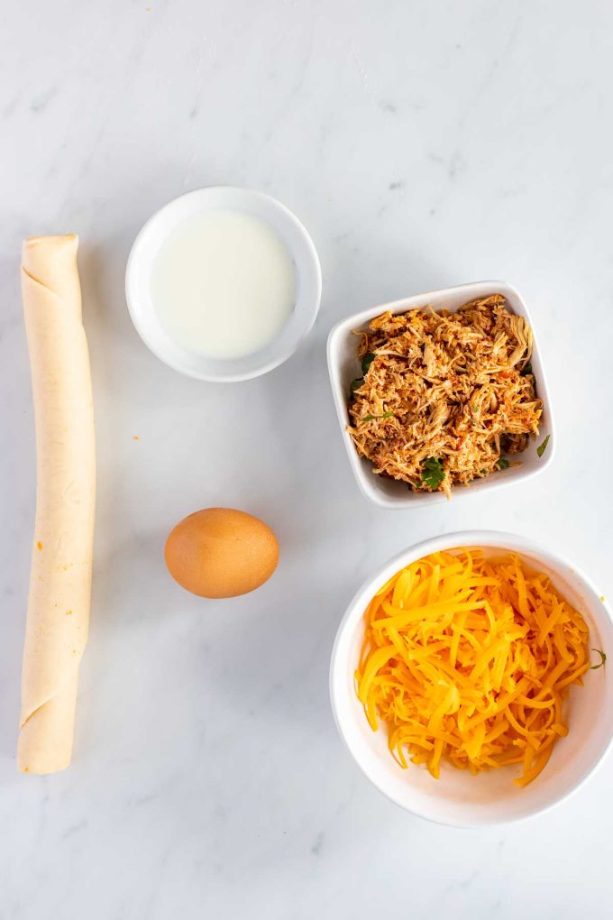 Overhead photo of a pie crust, egg, bowl of shredded cheese, bowl of shredded chicken and small dish of milk.