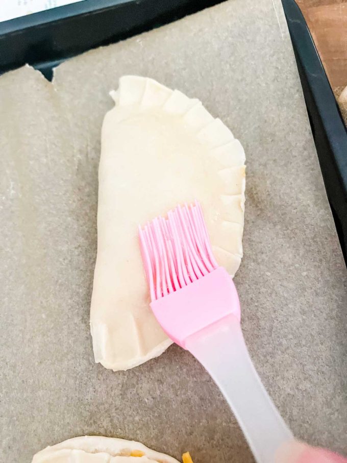 Photo of a pink silicone brush brushing an egg wash over an empanada.