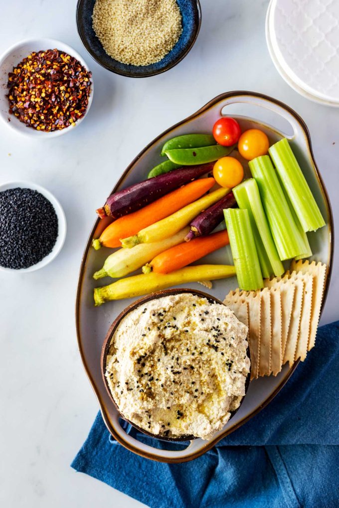 Overhead photo of a bowl of cauliflower hummus with crackers, celery, and carrots on a platter next to it and sesame seeds and crushed red pepper flakes on a platter next to it..
