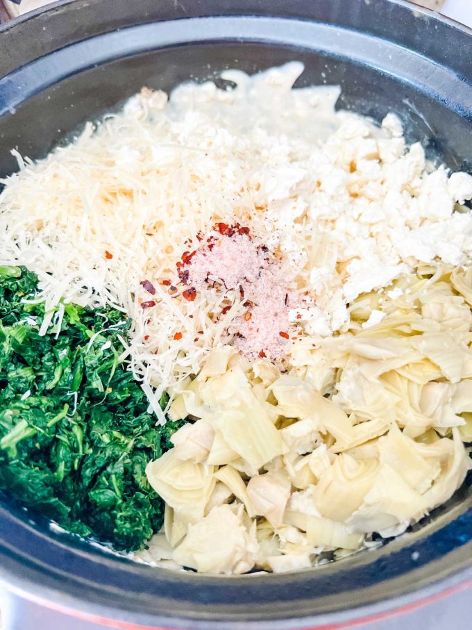 Photo of spinach, cream cheese, cheese, artichokes and seasonings being added to an slow cooker to make spinach artichoke dip.