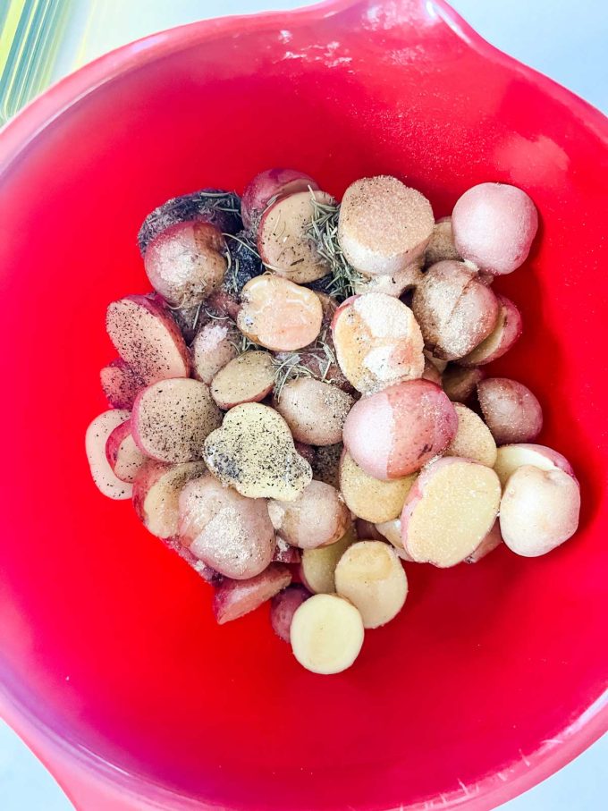 Photo of a red bowl of halved baby potatoes with seasonings on top.
