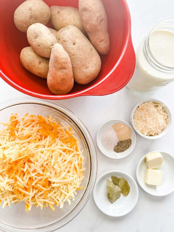 Overhead photo of a bowl of potatoes, shredded cheese, container of half and half, and prep bowls with butter, parmesan, seasonings, and bay leaves.