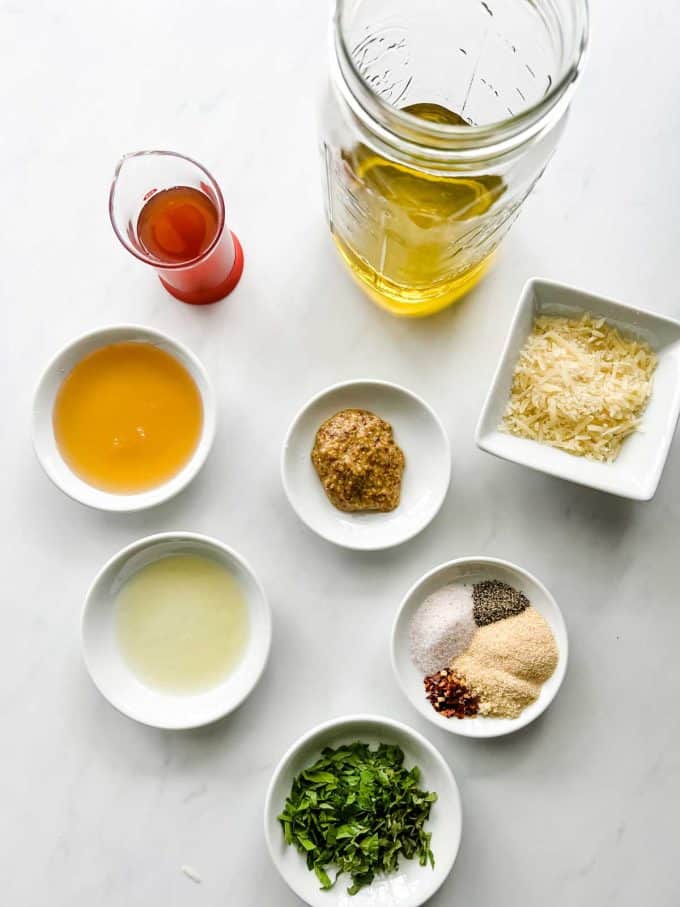 Photo of oil in a mason jar with small prep containers of vinegar, honey, mustard, parmesan, seasonings, lemon juice, and fresh basil.