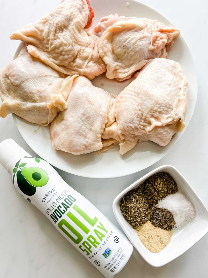 Photo of a plate of chicken thighs, a small dish of seasonings, and avocado oil spray.