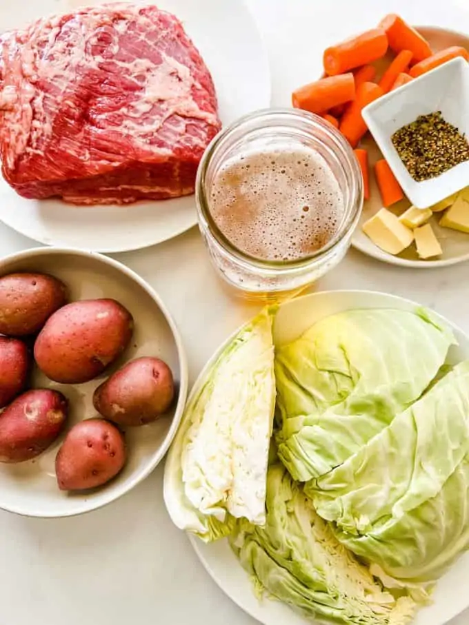 Overhead photo of corned beef, beer, potatoes, cabbage, carrots, beer, butter and seasonings in prep bowls.