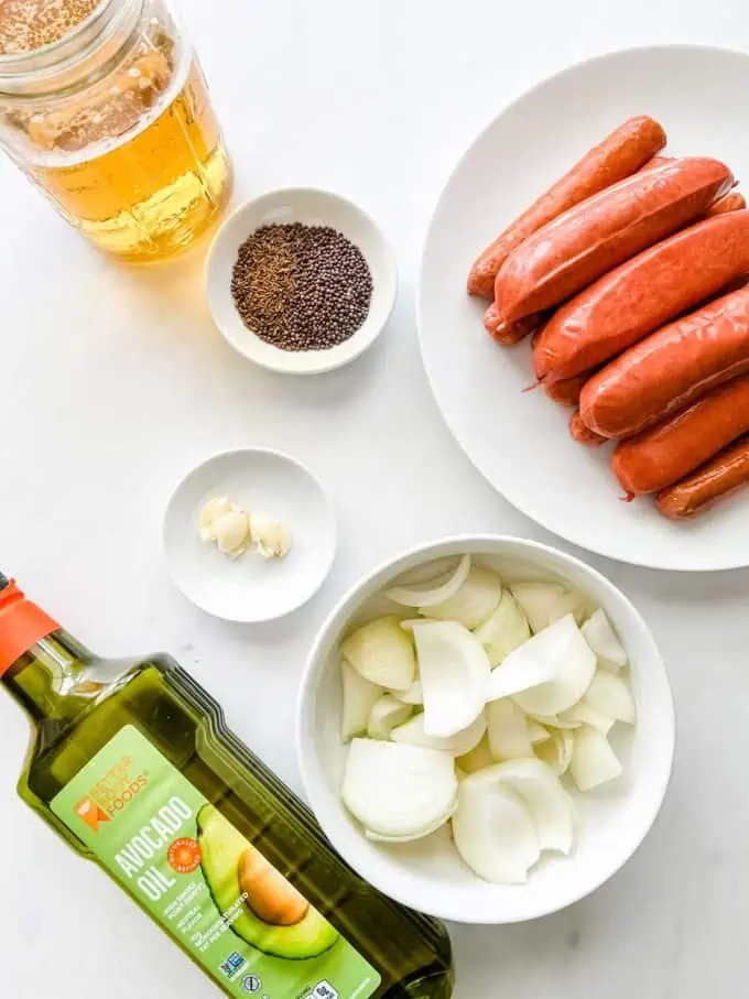 Photo of a mason jar of beer, bottle of avocado oil, plate of brats, onion, garlic, and seasonings.