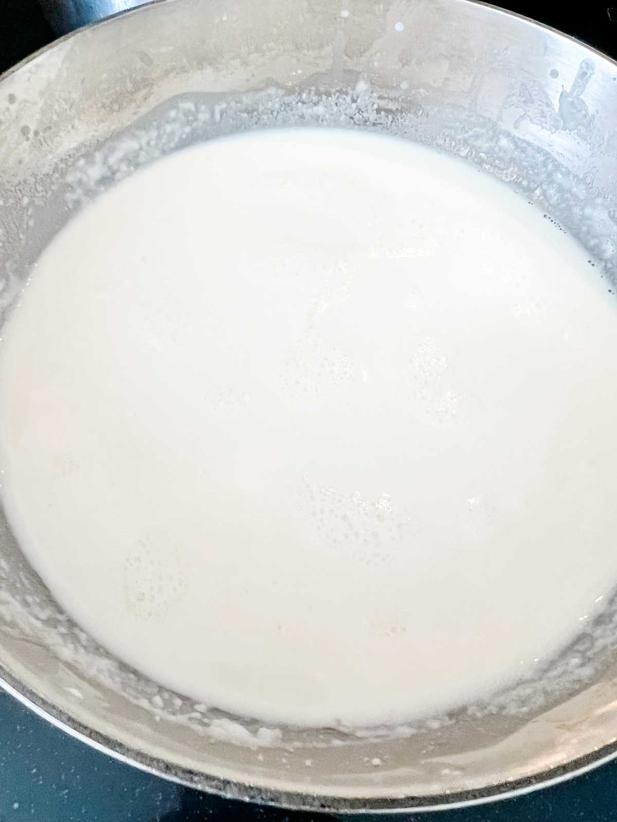 Photo of the remainder of the milk being added to a roux in a saucepan.