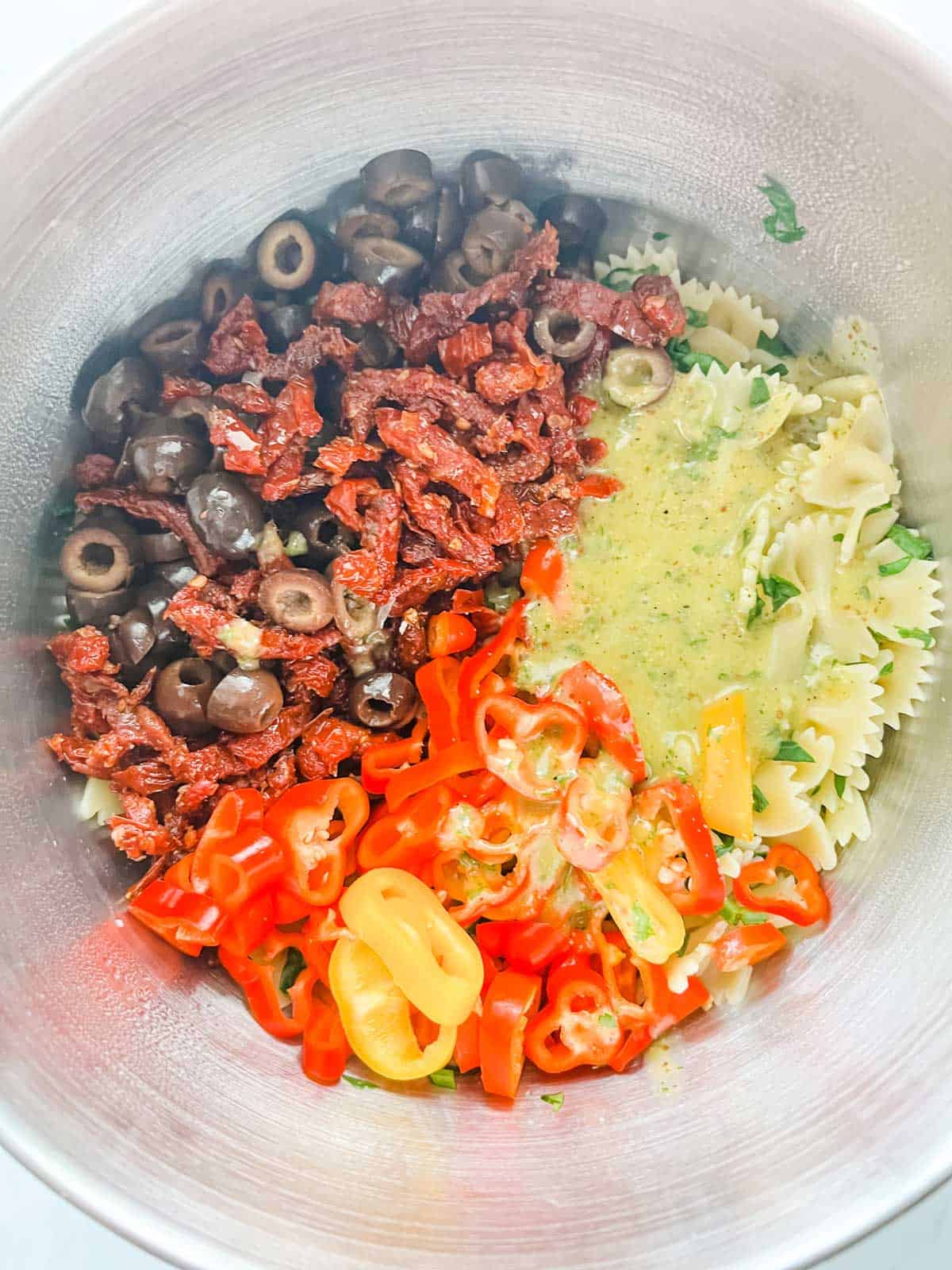 Cooked pasta, olives, sun dried tomatoes, sliced baby peppers and homemade salad dressing in a large bowl.