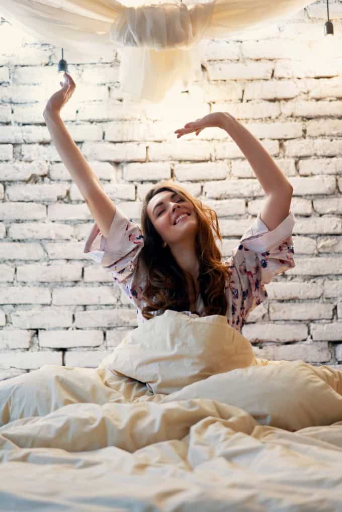 Photo of a woman waking up in a bed with a white brick wall in the background.