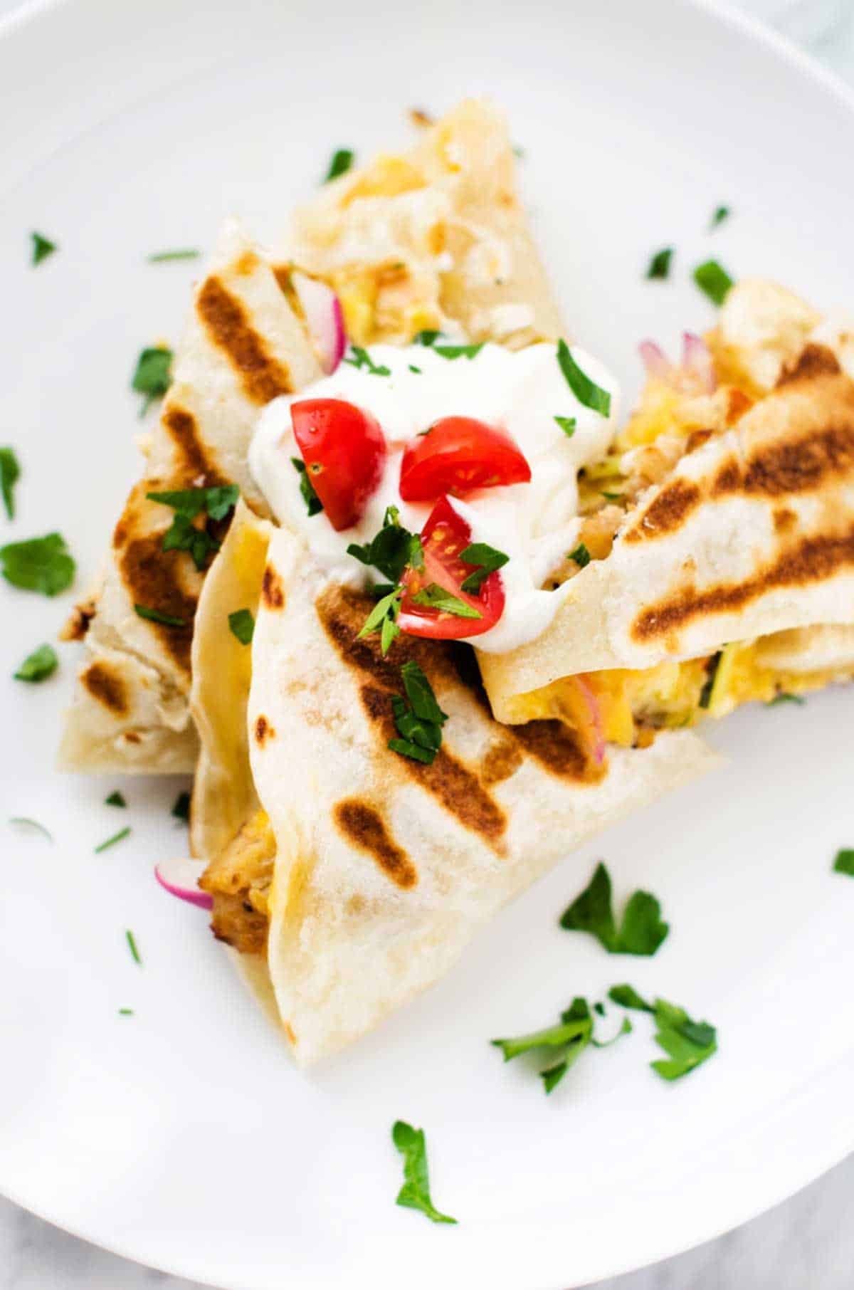 Close up photo of grilled quesadillas on a white plate garnished with sour cream, tomato and cilantro.