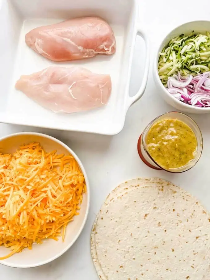 Overhead photo of chicken breast in a casserole dish, bowls of zucchini, red onion, and cheddar cheese, Italian dressing and tortillas.