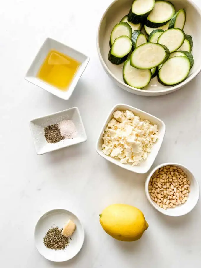 Overhead photo of sliced zucchini oil, salt, pepper, feta, pine nuts, lemon, thyme, and garlic in prep dishes.
