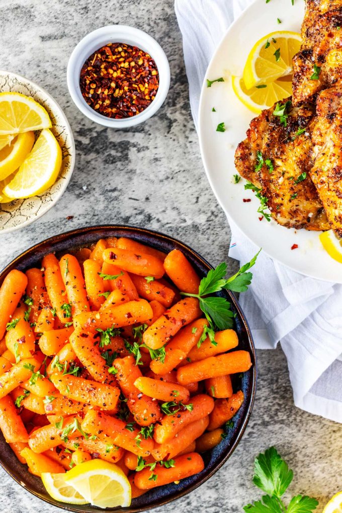 Overhead photo of a bowl of Instant Pot Baby Carrots sitting next to small dishes of slice lemons and crushed red pepper flakes and a platter of chicken thighs.