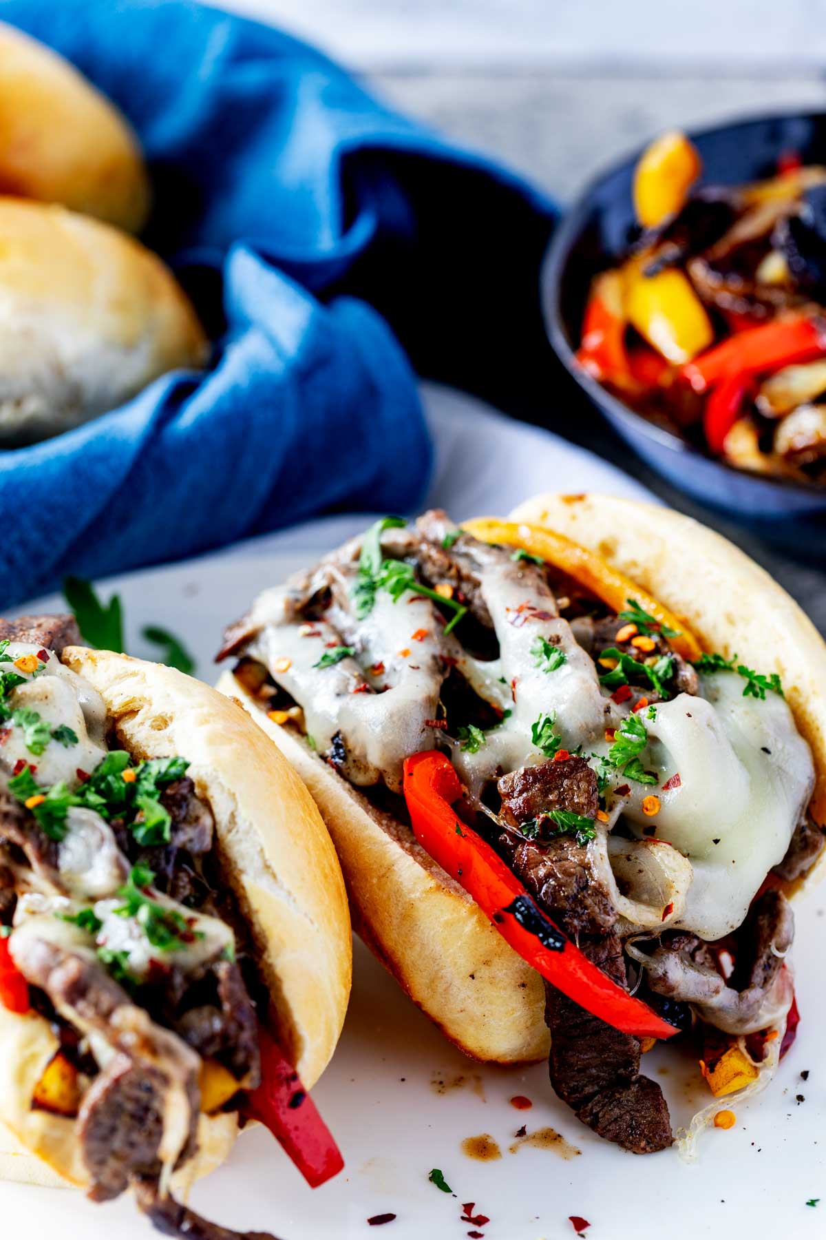 Photo of a plate of Blackstone Philly Cheesesteak with a bowl of peppers and onions and basket of buns behind it.