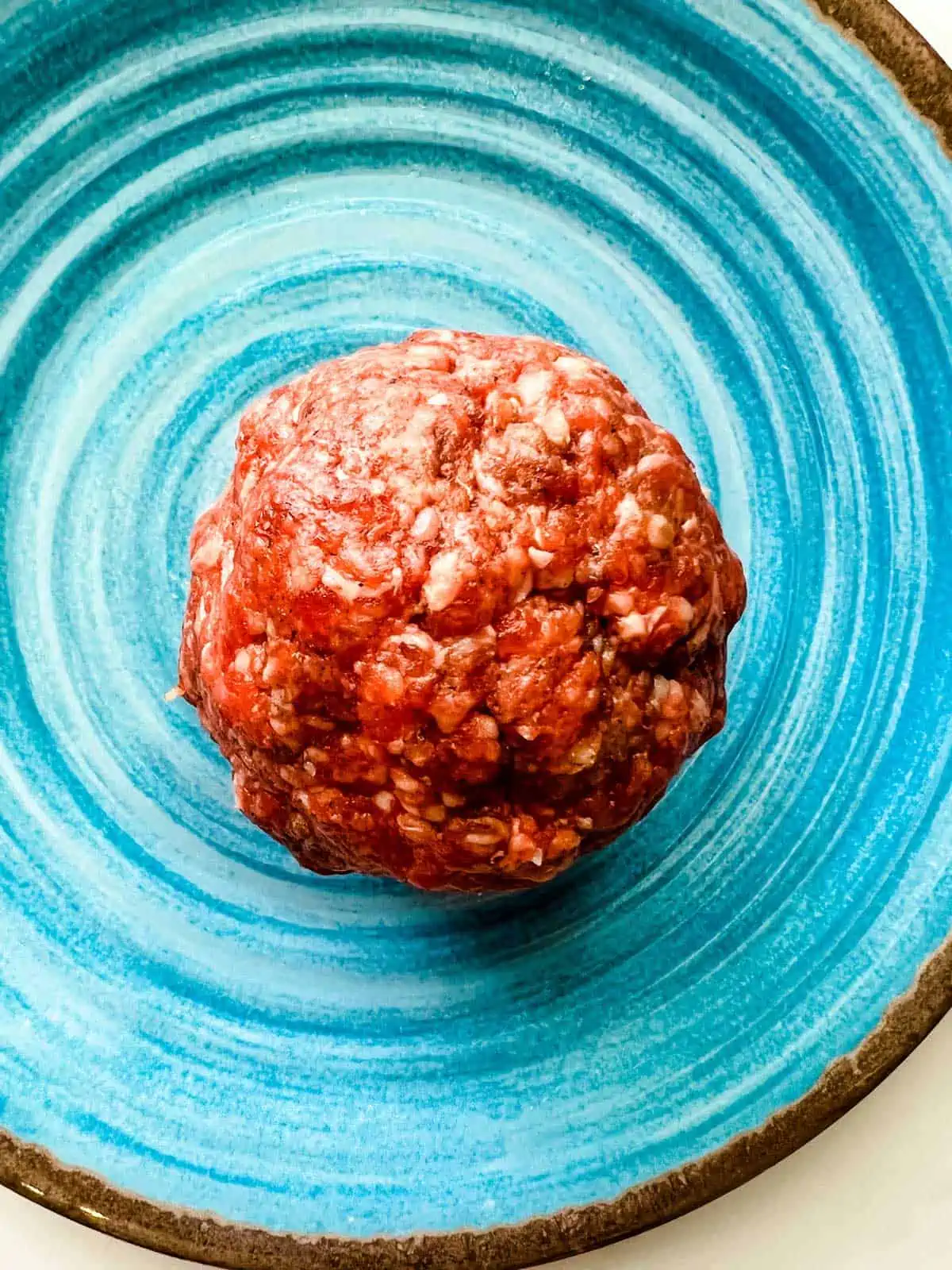 Photo of a ball of of seasoned ground beef to make a burger on a blue plate.