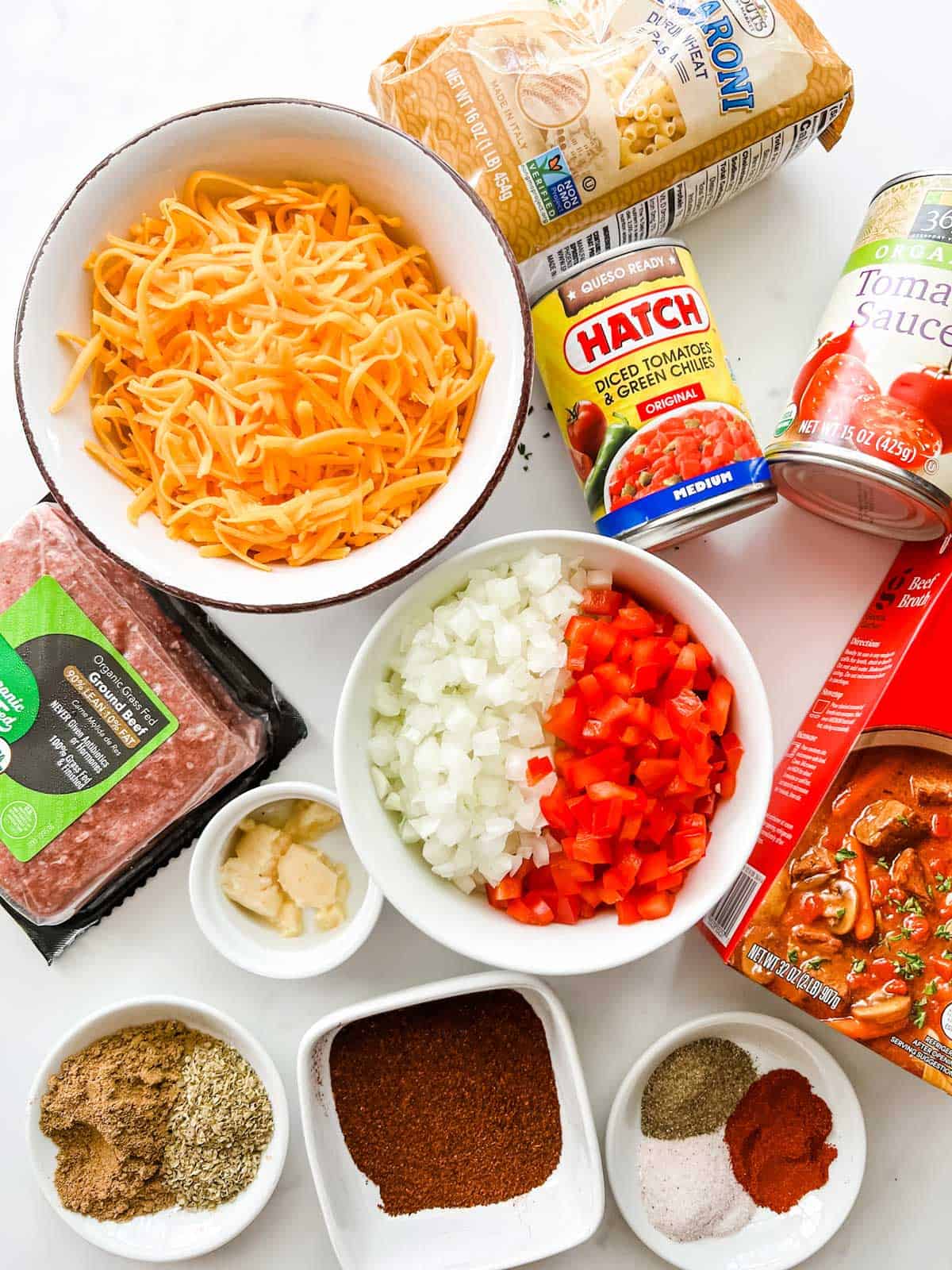 Overhead photo of prepped ground beef, onion, red pepper, garlic, chili powder, cumin, oregano, salt, paprika, black pepper, beef broth, tomatoes, tomato sauce, elbow noodles, and shredded cheese.