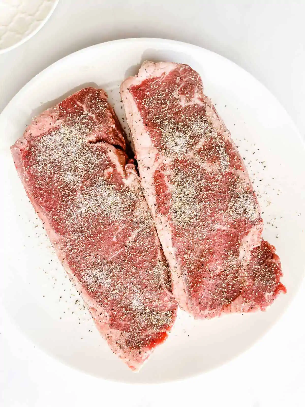 Steaks that have been seasoned sitting on a white plate.