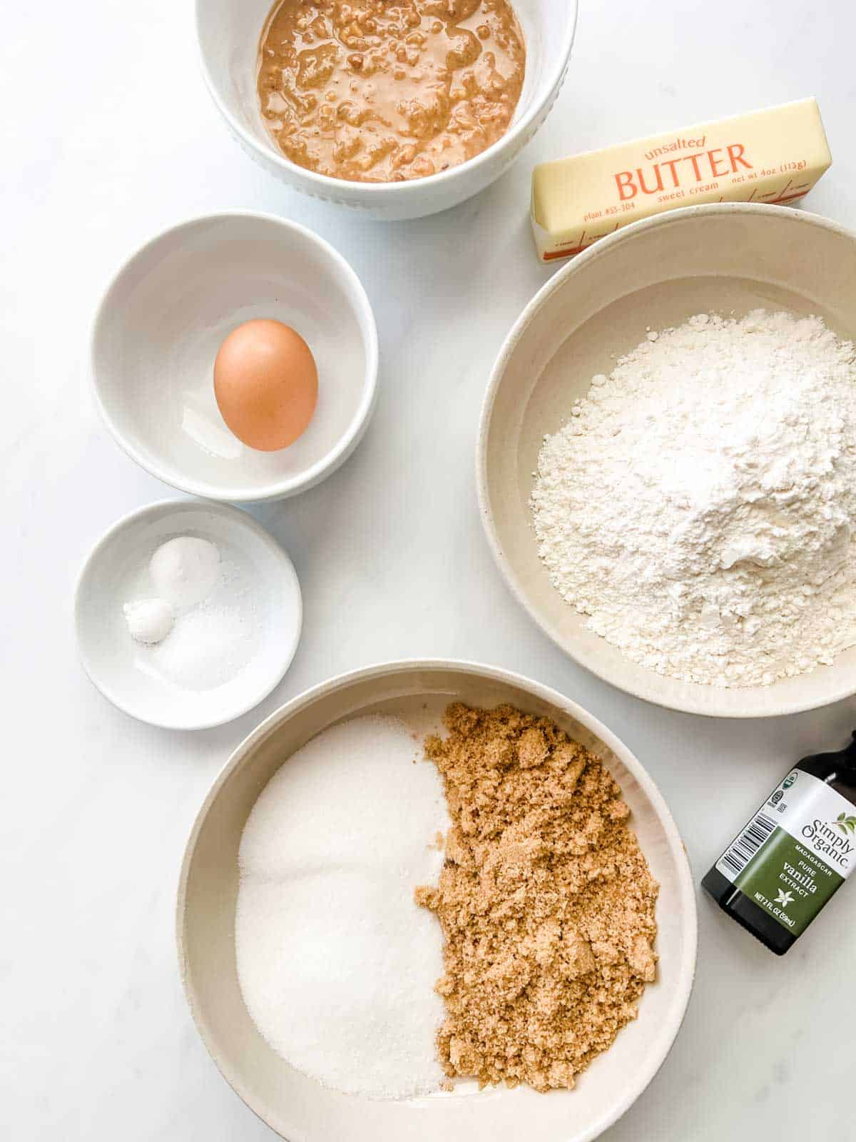 Overhead photo of prep bowls with peanut butter, an egg, baking powder, white sugar, brown sugar, flour, vanilla, and butter.