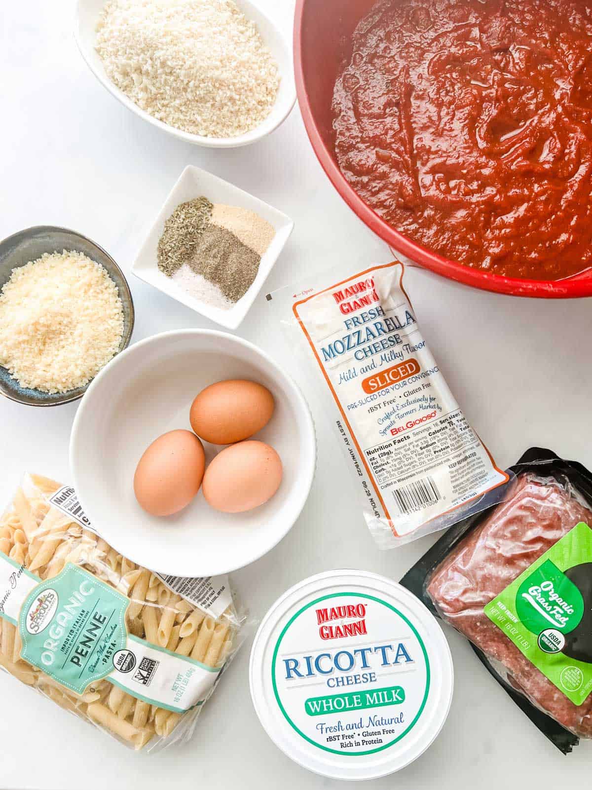 Overhead photo of the ingredients for meatball casserole: marinara, ground beef, mozzarella, ricotta, eggs, penne, parmesan, breadcrumbs, and seasonings.