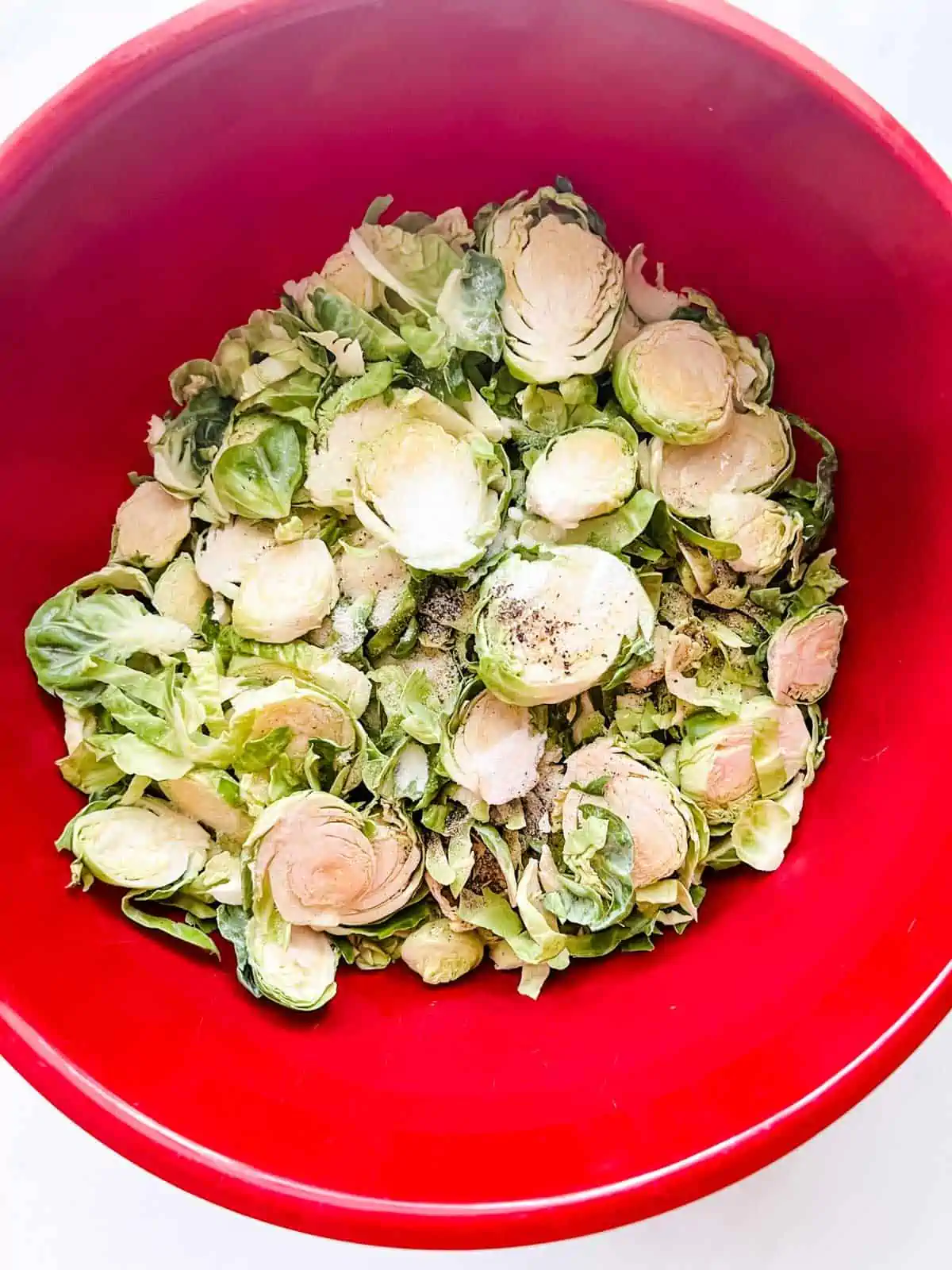 Photo of a red bowl with shaved brussels sprouts, seasonings, and oil.