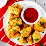 Square overhead photo of a white plate with air fryer chicken nuggets and ketchup sitting on a red and white napkin.