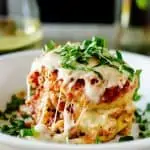 Square side photo of a white plate of eggplant parmesan sitting on a wooden background and garnished with fresh basil.