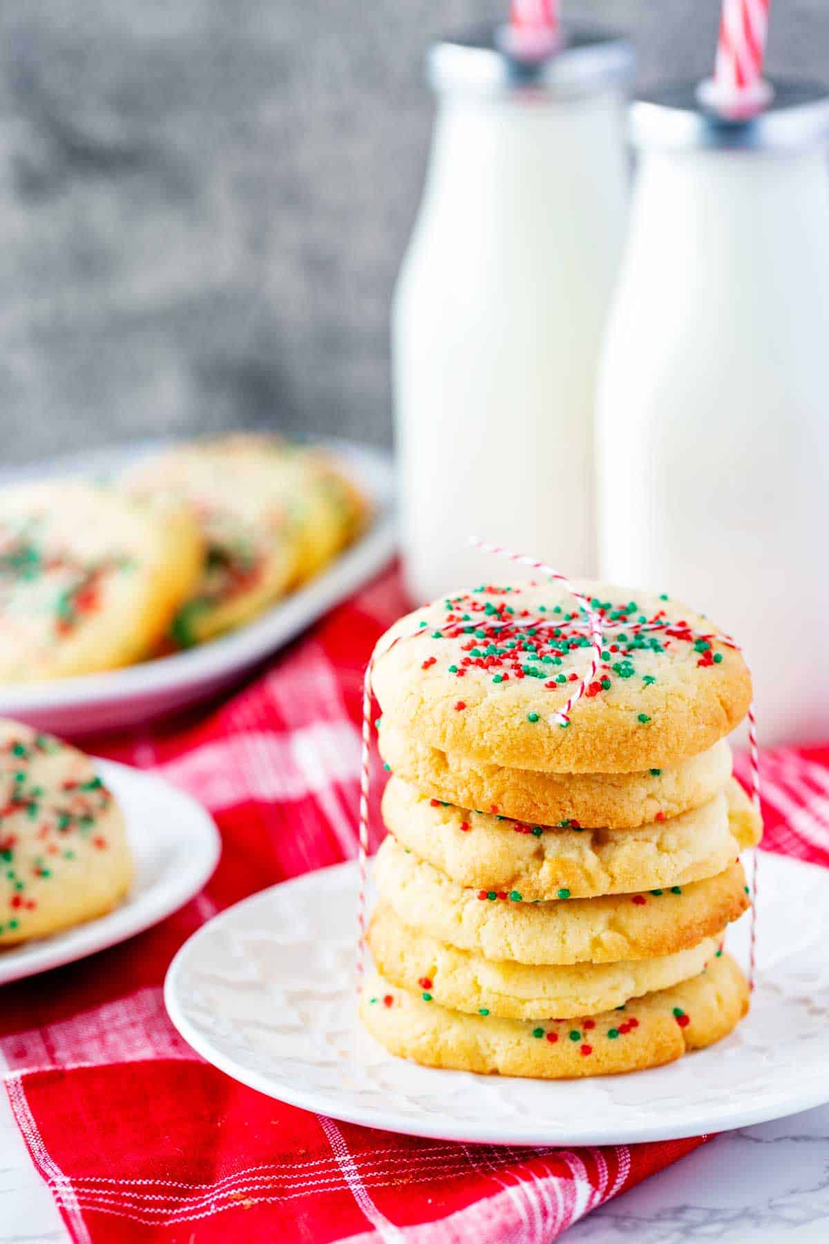 Photo of a plate of stacked air fryer sugar cookies with two other plates of cookies and milk bottles around it.