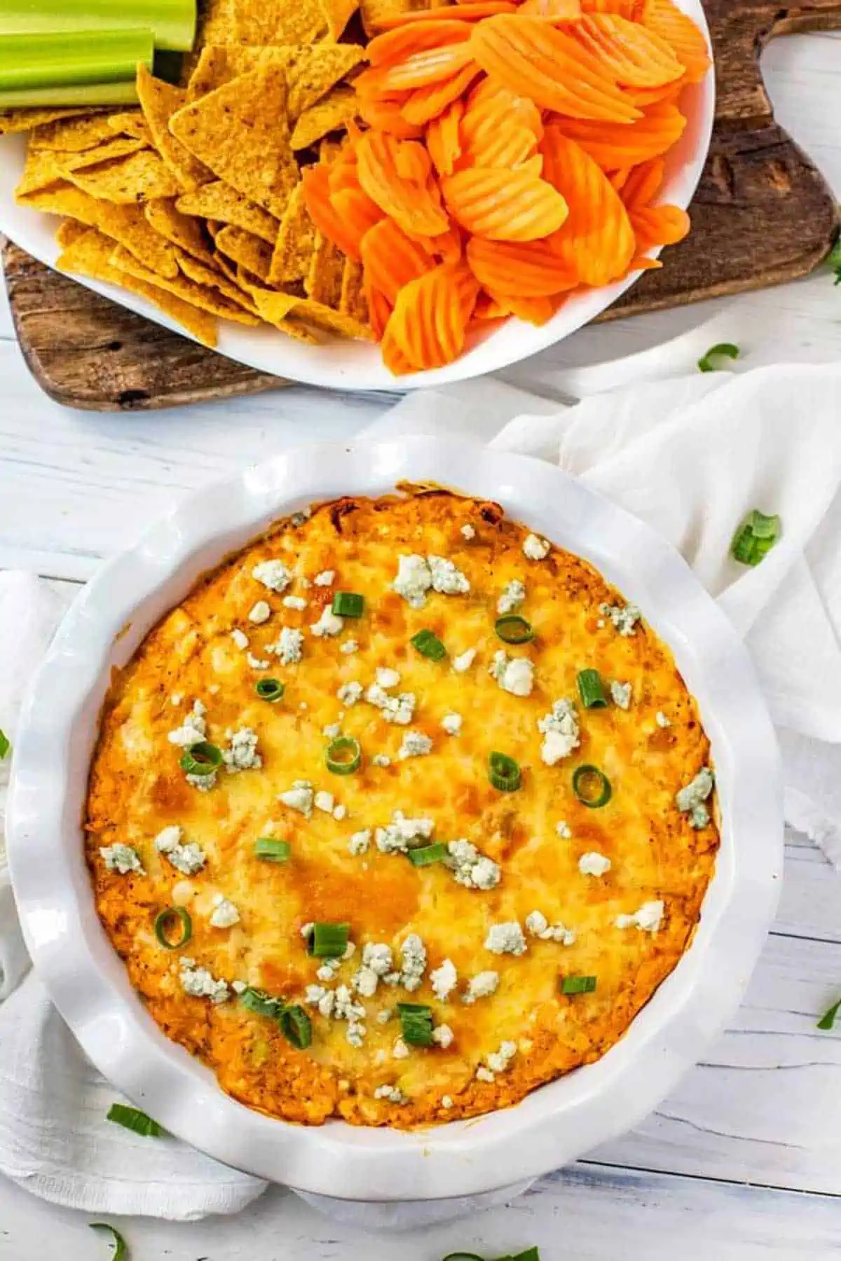 Photo of a Healthy Buffalo Chicken Dip in a white pie plate garnished with blue cheese and scallions. Another plate of carrots, celery and chips are behind it.