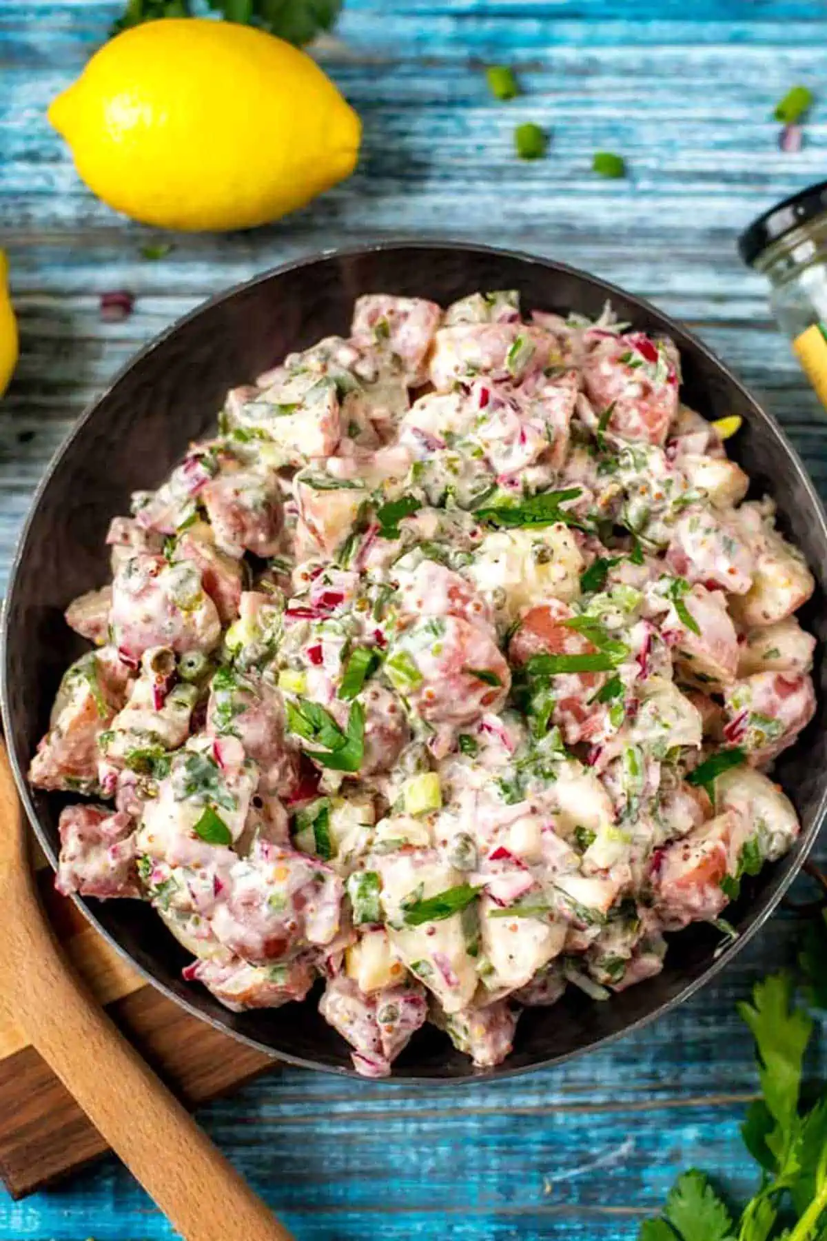 A dark bowl with red skin potato salad surrounded by lemon, herbs, and a wooden serving spoon.