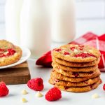 Square side photo of a stack of white chocolate raspberry cookies with raspberries and chocolate chips beside it and jars of milk behind it.