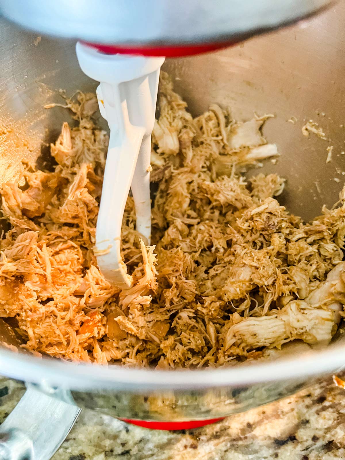 Chicken being shredded in the bowl of a stand mixer.