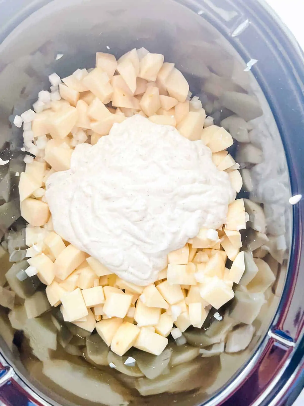 Photo of a creamy mixture that has been poured over diced onion and potatoes in a slow cooker.