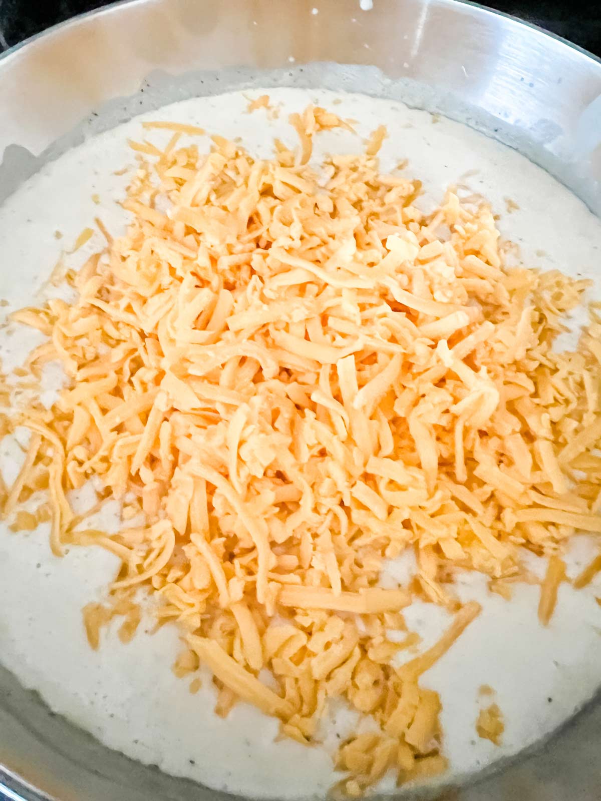 A saucepan with cream sauce that has just had shredded cheddar sprinkled over it.